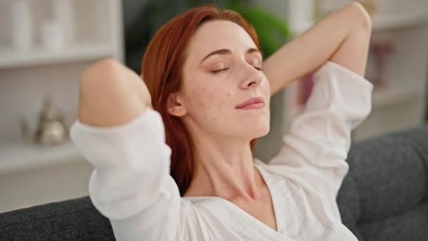 Giovane donna rossa rilassata con le mani sulla testa seduta sul divano a casa - Filmati, video