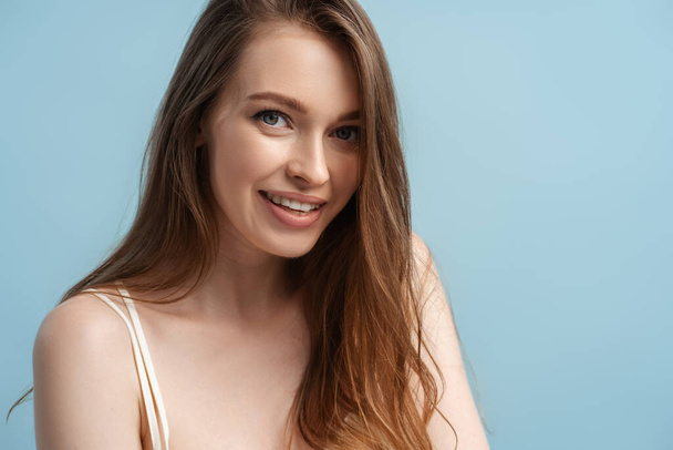 Schönheitsporträt einer jungen lächelnden Frau mit langen Haaren, die isoliert in die Kamera auf blauem Hintergrund blickt. Natürliche Schönheit, Haarpflege, Morgenroutine-Konzept  - Foto, Bild