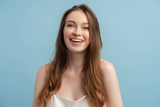 Auténtico retrato de joven hermosa mujer sonriente con pelo brillante, maquillaje natural con vestido elegante aislado sobre fondo azul  - Foto, imagen