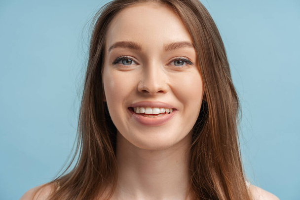 Close-up portret mooie glimlachende vrouw met tanden glimlach op zoek naar camera na tandheelkundige ingreep. Aantrekkelijk modemodel met natuurlijke make-up op het gezicht geïsoleerd op blauwe achtergrond. Natuurlijke schoonheid - Foto, afbeelding