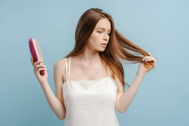 Portrait of worried beautiful woman in nightgown holding hairbrush, isolated on blue background. Young female with hair loss. ?oncept of treatment, health care - Photo, Image