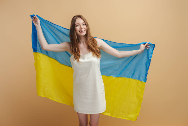Portré gyönyörű mosolygós nő elegáns ruhában fogas mosollyal gazdaság ukrán zászló, elszigetelt bézs háttérrel. Vonzó fiatal nő pózol a stúdióban, kamerába néz - Fotó, kép