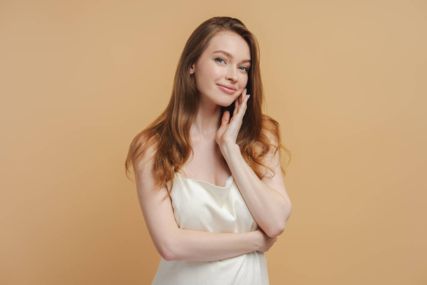 Portret van aantrekkelijke mooie vrouw in jurk of nachtjapon geïsoleerd op beige achtergrond. Glimlachende vrouw met mooi lang haar die alleen naar de camera kijkt. Natuurlijke schoonheid concept - Foto, afbeelding