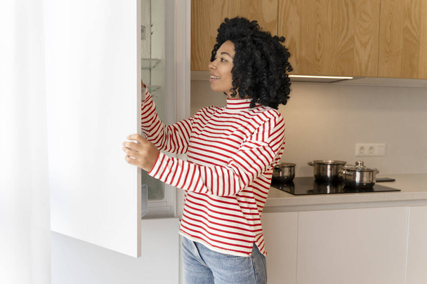 Linda, atraente mulher afro-americana que abre a geladeira na cozinha moderna elegante. Dona de casa encaracolada usando eletrodomésticos. Conceito de equipamento, doméstico - Foto, Imagem