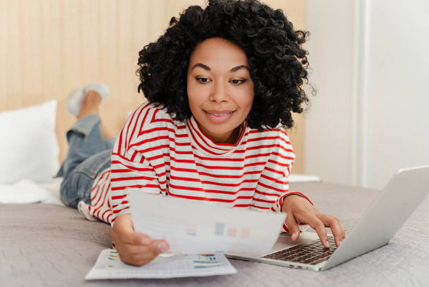 Portret van een lachende aantrekkelijke Afro-Amerikaanse vrouw op bed in de woonkamer met behulp van een laptop, werken met grafieken, documenten. Curly stijlvolle hipster werken online. Concept van werk op afstand - Foto, afbeelding