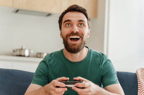 Portret van een opgewonden knappe bebaarde mannelijke gamer die videospel speelt, joystick vasthoudt, op een comfortabele bank zit in de woonkamer thuis, close-up. Glimlachende man draagt casual kleding online plezier hebben - Foto, afbeelding
