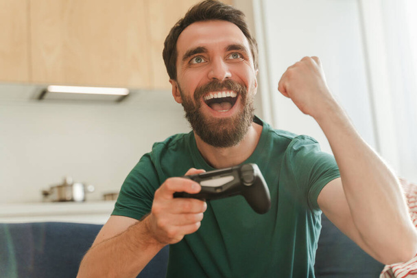Emocionado hombre sonriente sentado en cómodo sofá en la cocina en el apartamento haciendo gesto ganador jugando videojuegos. Jugador masculino positivo con barba divirtiéndose. Concepto tecnológico - Foto, Imagen