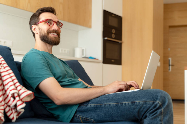 Улыбающийся бородатый мужчина, программист в очках с помощью ноутбука, работающего онлайн из дома - Фото, изображение