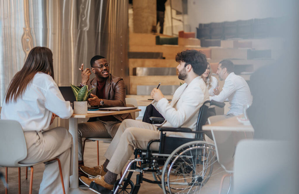 Homme attrayant en fauteuil roulant portant un costume blanc assis avec ses amis tout en faisant la conversation sur le plan d'affaires - Photo, image