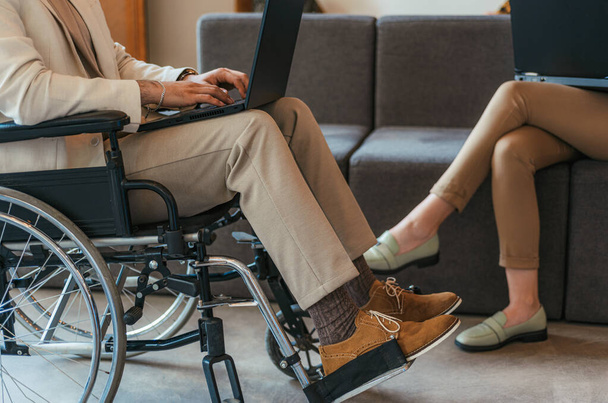 Niepełnosprawny mężczyzna na wózku inwalidzkim. Pracuję nad okrążeniem. Kobieta siedząca na kanapie i pracująca na kolanach. Przycięte zdjęcie - Zdjęcie, obraz