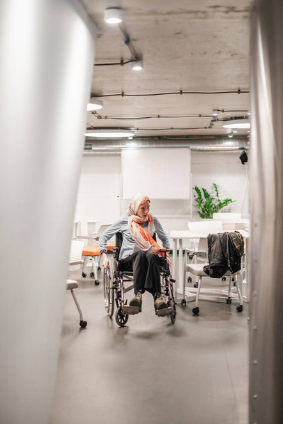 Μια γυναίκα με ειδικές ανάγκες κοιτάζει κάτι ενώ κυλά την αναπηρική καρέκλα της στην αίθουσα συνεδριάσεων. - Φωτογραφία, εικόνα