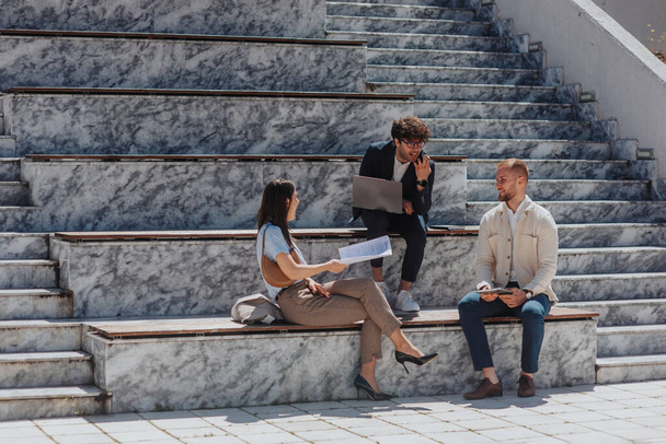 Όμορφοι νέοι επιχειρηματίες κάθονται στις σκάλες της πόλης, εργάζονται μαζί, διασκεδάζουν και χαμογελούν - Φωτογραφία, εικόνα