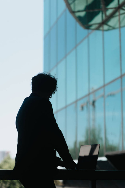 Man with wavy hair working on lap top placed on a metal fence in front of big corporate building. Silhouette photo - Photo, Image