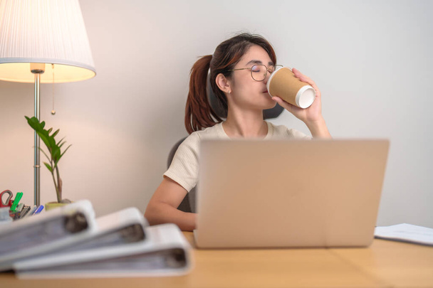 Στρεσαρισμένη Ασιάτισσα γυναίκα που πίνει καφέ ενώ εργάζεται, γυναίκα επιχειρηματίας που χρησιμοποιεί φορητό υπολογιστή με κουρασμένο και υπνηλία στο γραφείο, εξαντλημένη γυναίκα με υπολογιστή στο σπίτι αργά το βράδυ. Υπερφόρτωση και υπερκόπωση - Φωτογραφία, εικόνα