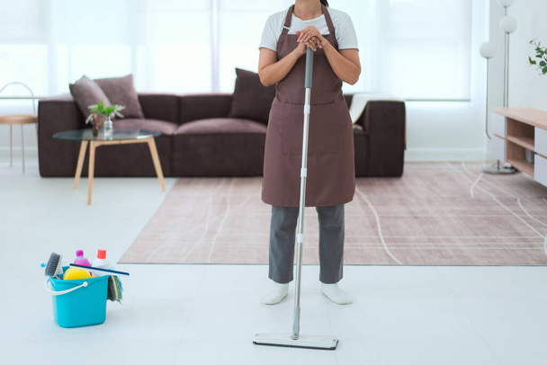 Schoonmaakster staat in de woonkamer en houdt dweil met emmer plastic van schonere apparatuur op de vloer. - Foto, afbeelding