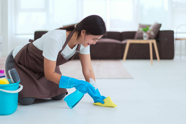 Pokojówka nosi rękawiczki i używa czystszego sprayu i ściereczki z mikrofibry do wycierania kurzu na podłodze.. - Zdjęcie, obraz