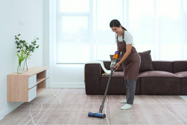 Maid holds wireless cordless vacuum cleaner to vacuuming dust and cleaning on carpet in living room. - Photo, Image
