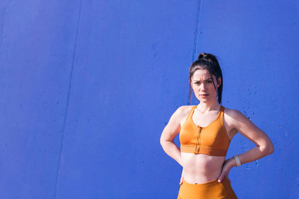portrait of a serious sportswoman posing with hands on hips looking at the camera in front of a blue wall, concept of sport and active lifestyle, copy space for text - Foto, Imagem