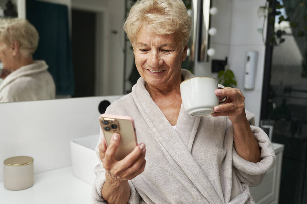 Seniorin trinkt Kaffee und telefoniert im Badezimmer - Foto, Bild