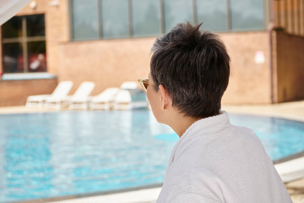femme mature en lunettes de soleil et peignoir reposant dans le pavillon privé près de la piscine pendant les vacances - Photo, image