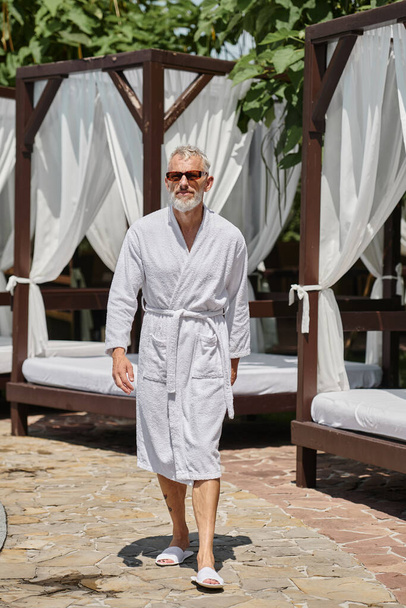 мужчина средних лет в стильных солнцезащитных очках и белом халате, идущий рядом с частным павильоном в роскошном курорте - Фото, изображение