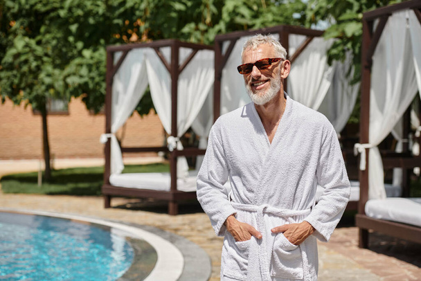 hombre maduro alegre en gafas de sol y bata posando con las manos en bolsillos cerca de la piscina en el resort - Foto, Imagen
