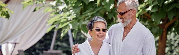 pareja madura feliz en gafas de sol y batas en resort de lujo, concepto de retiro de bienestar, bandera - Foto, Imagen