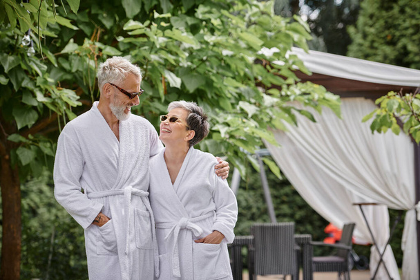 alegre hombre maduro con tatuaje abrazando esposa en gafas de sol y bata, jardín de verano, retiro de bienestar - Foto, Imagen