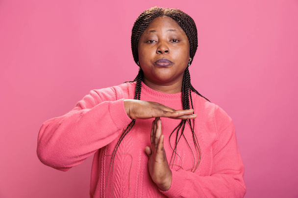 Разгневанная женщина делает тихий жест в форме футболки, делая знак времени перерыва руками в студии на розовом фоне. Африканский американский молодой взрослый рекламный символ паузы с ладонями - Фото, изображение