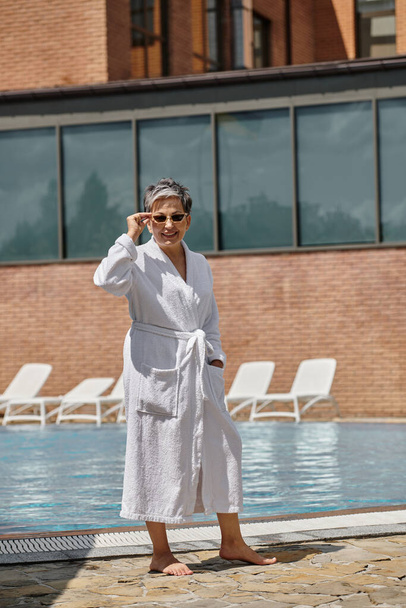 高級リゾートのプールサイドに立っている白いローブとサングラスの陽気な中年女性 - 写真・画像