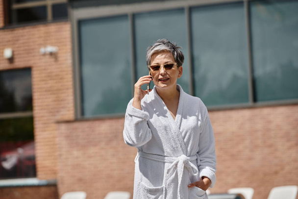 жінка середнього віку в білому халаті і сонцезахисних окулярах розмовляє на смартфоні біля басейну в розкішному курорті - Фото, зображення