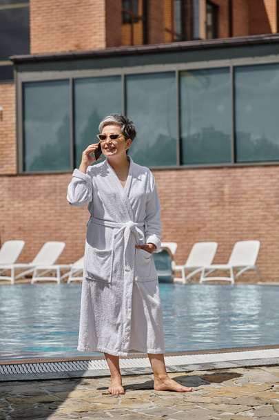 счастливая женщина средних лет в халате и солнцезащитных очках разговаривает на смартфоне у бассейна в роскошном курорте - Фото, изображение