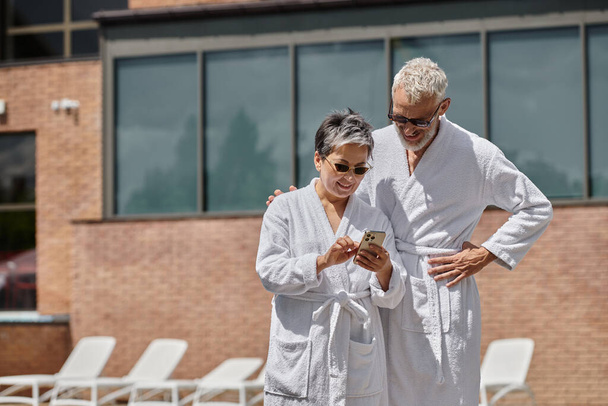 szczęśliwa kobieta w średnim wieku w okularach przeciwsłonecznych za pomocą smartfona w pobliżu męża na basenie w luksusowym kurorcie - Zdjęcie, obraz