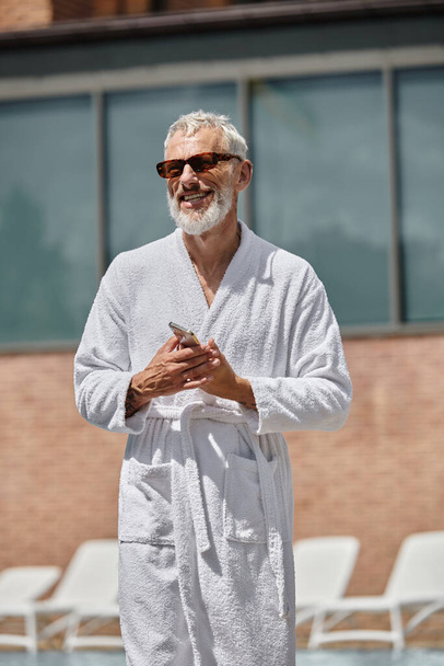 hombre de mediana edad feliz y tatuado en gafas de sol y bata con teléfono inteligente en el resort, retiro - Foto, imagen