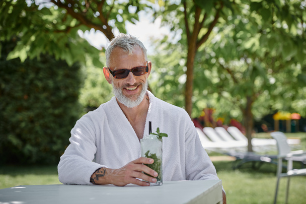 Wellness-Rückzugskonzept, glücklicher Mann mittleren Alters in Sonnenbrille und Bademantel genießt Cocktail im Urlaub - Foto, Bild