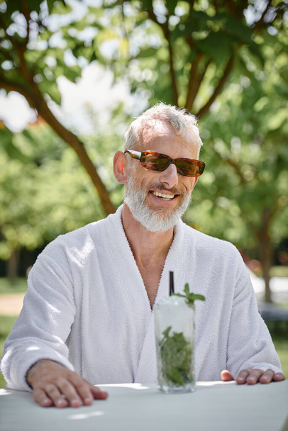 Konzept des Wellness-Rückzugs, glücklicher reifer Mann in Sonnenbrille und Robe beim Cocktail im Urlaub - Foto, Bild