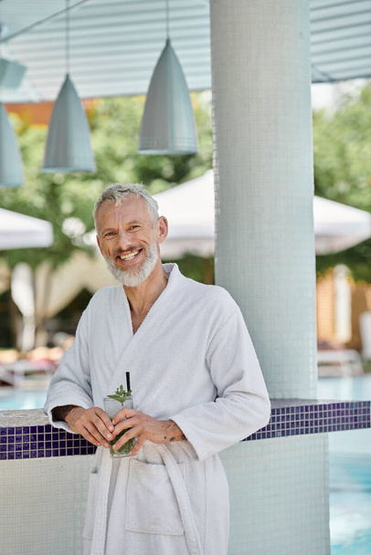 Mann mittleren Alters in weißer Robe lächelt und hält Mojito-Cocktail am Pool, Wellness-Rückzugsort - Foto, Bild