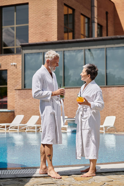 fröhliches reifes Paar in weißen Roben mit Cocktails am Pool, Wellness-Rückzugsort - Foto, Bild