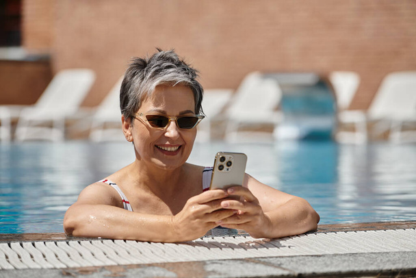 femme d'âge moyen gaie dans des lunettes de soleil en utilisant smartphone à l'intérieur de la piscine, retraite bien-être - Photo, image