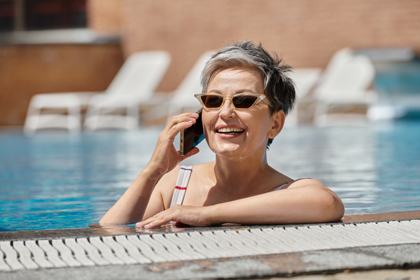 šťastný dospělý žena v slunečních brýlích mluví na smartphone uvnitř bazénu, wellness ústup - Fotografie, Obrázek