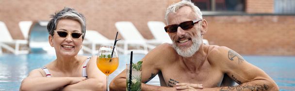 счастливая зрелая пара в солнцезащитных очках, купающаяся в бассейне, улыбающаяся рядом с коктейлями, оздоровительный отдых, баннер - Фото, изображение