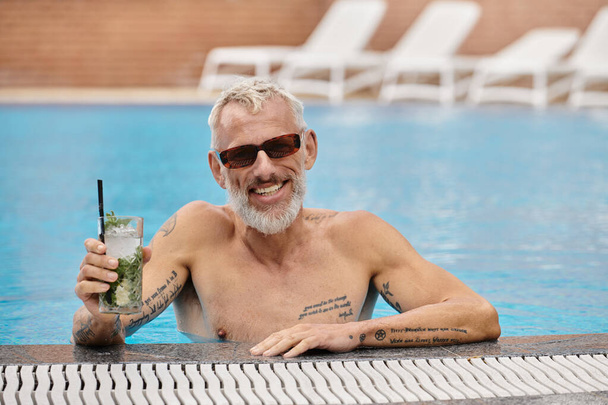 shirtloze en getatoeëerde man van middelbare leeftijd in een zonnebril met cocktail en zwemmen in het zwembad, retraite - Foto, afbeelding