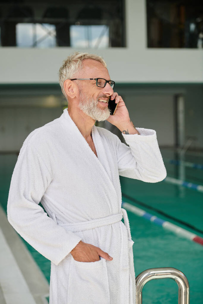 hombre maduro alegre en bata blanca y gafas hablando en el teléfono inteligente dentro del centro de spa, retiro - Foto, imagen