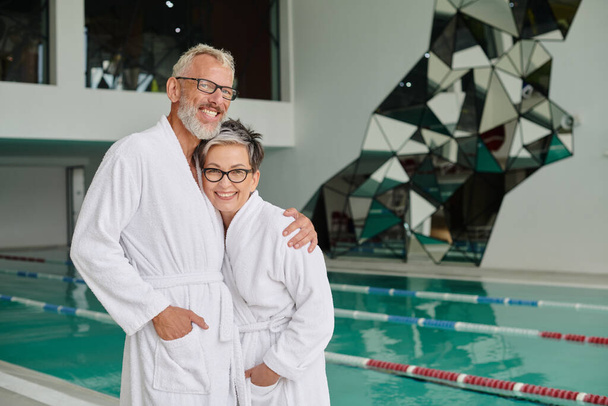 feliz homem de meia idade em óculos e branco esposa roupão abraçando no centro de spa, conceito de retiro de bem-estar - Foto, Imagem