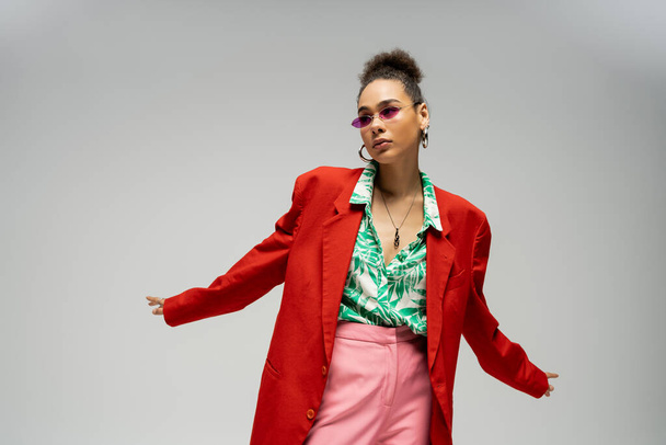 stylish african american woman in  sunglasses and trendy vibrant attire posing on grey backdrop - Photo, Image