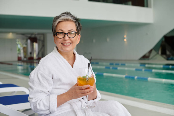 spa center concept, happy mature woman in glasses and robe holding cocktail and sitting on lounger - Photo, Image