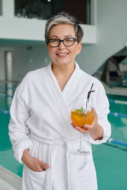 spa and retreat concept, happy mature woman in glasses and robe holding cocktail near indoor pool - 写真・画像
