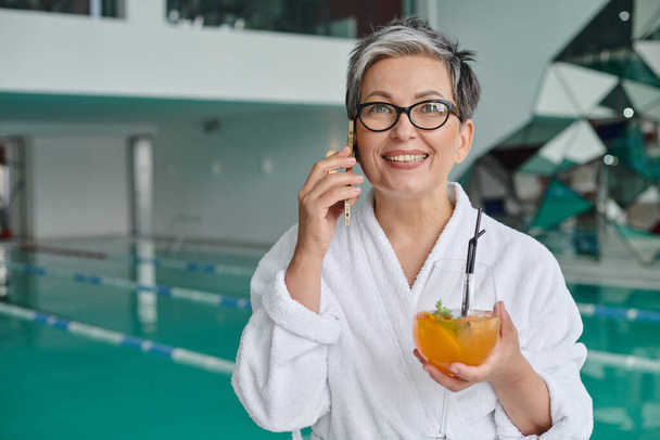 retreat concept, happy mature woman in glasses and robe holding cocktail and talking on smartphone - Photo, Image