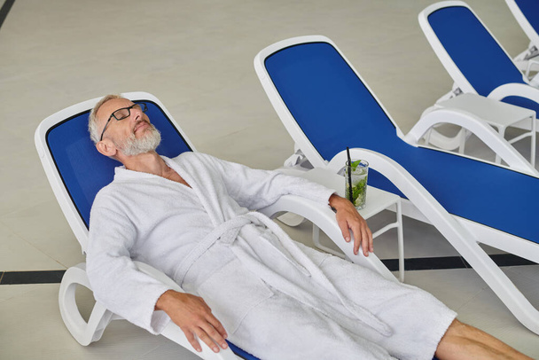 concept de bien-être, homme mature en lunettes et peignoir reposant sur une chaise longue près d'un cocktail dans un centre de spa - Photo, image