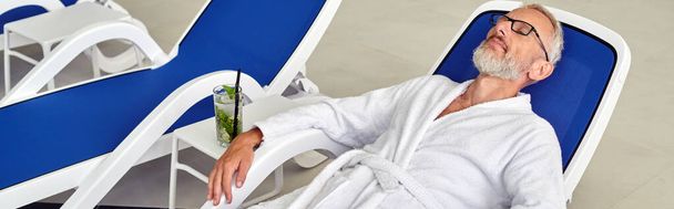 wellness concept, mature man in glasses and robe resting on lounger near cocktail in spa, banner - Photo, Image